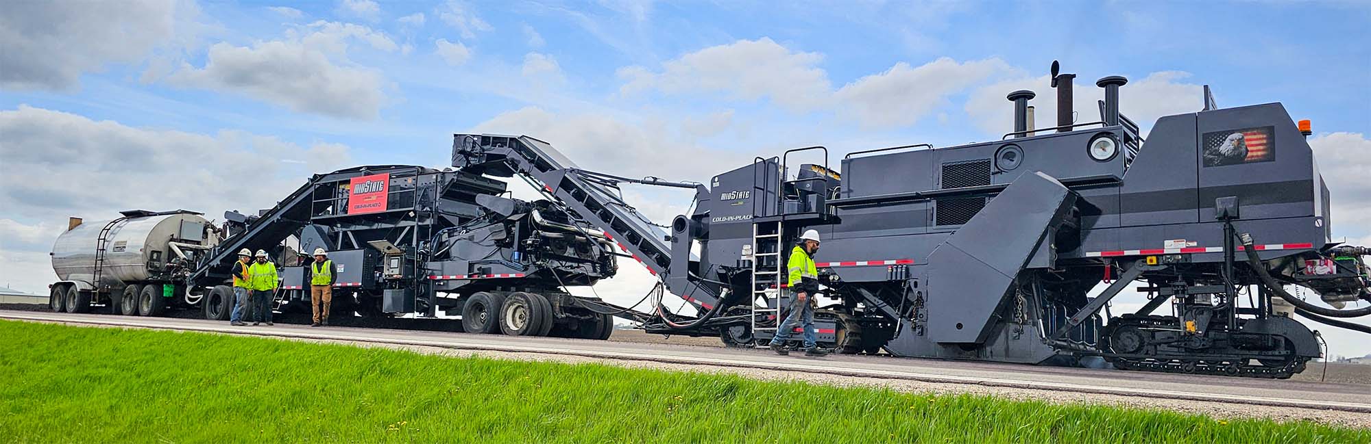 Cold In-Place Recycling (CIR) train working on a road construction site, with grassy fields and cloudy skies.